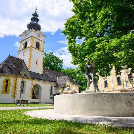 Eine Kirche mit Glockenturm und Statue steht in einem Grasbereich mit einem Baum, sonnigem Wetter und einem gelben Gebäude dahinter.