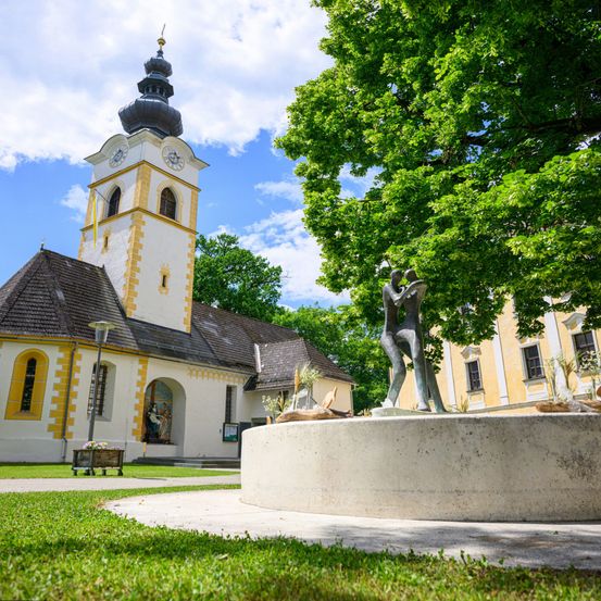 Eine Kirche mit Glockenturm und Statue steht in einem Grasbereich mit einem Baum, sonnigem Wetter und einem gelben Gebäude dahinter.