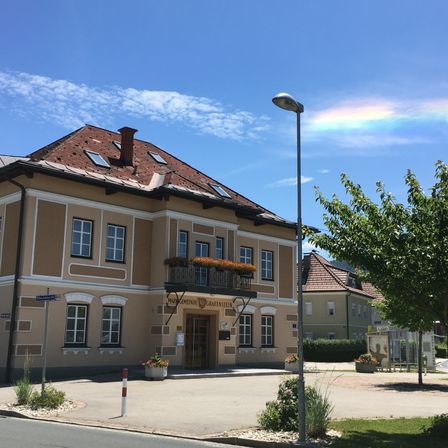 Ein Rathausgebäude mit Solarpanelen auf dem Dach. Es hat viele Fenster und einen Balkon mit Blumenvasen. Das Gebäude ist von Pflanzen und einem Baum umgeben. Eine Straßenlaterne steht am Straßenrand.