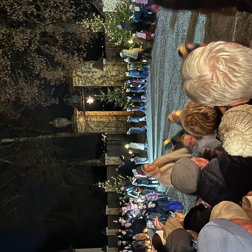 Eine große Menschenmenge versammelt sich im Freien, möglicherweise für ein Winterfest, mit warm gekleideten Menschen. Bäume mit Lichtern und ein Gebäude im Hintergrund.