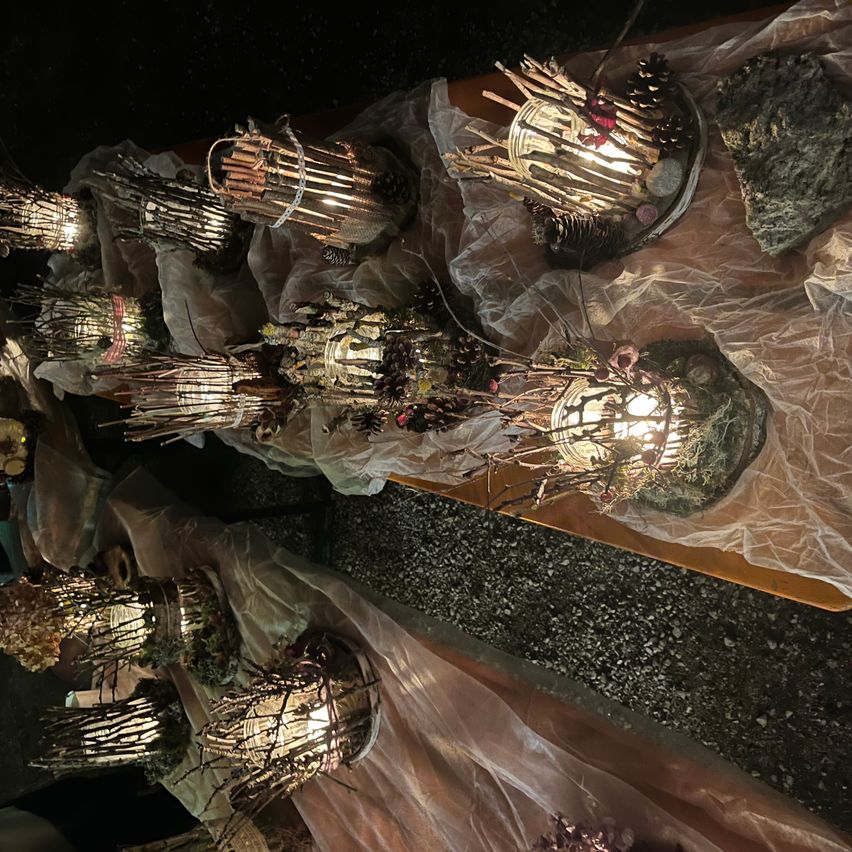 Eine Sammlung handgefertigter Laternen aus Zweigen und Steinen, jede mit einem warmen Leuchten, auf einem mit Plastik bedeckten Tisch ausgestellt.