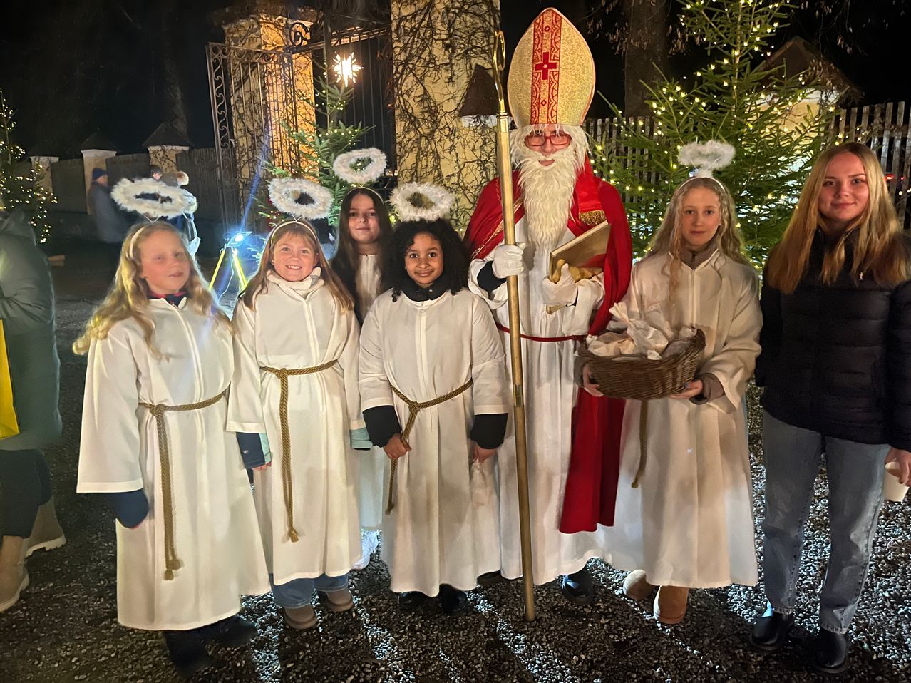 Fünf Kinder in weißen Gewändern mit Engelsflügeln stehen neben einem Mann als St. Nikolaus verkleidet, der einen Korb hält, vor einem beleuchteten Weihnachtsbaum.