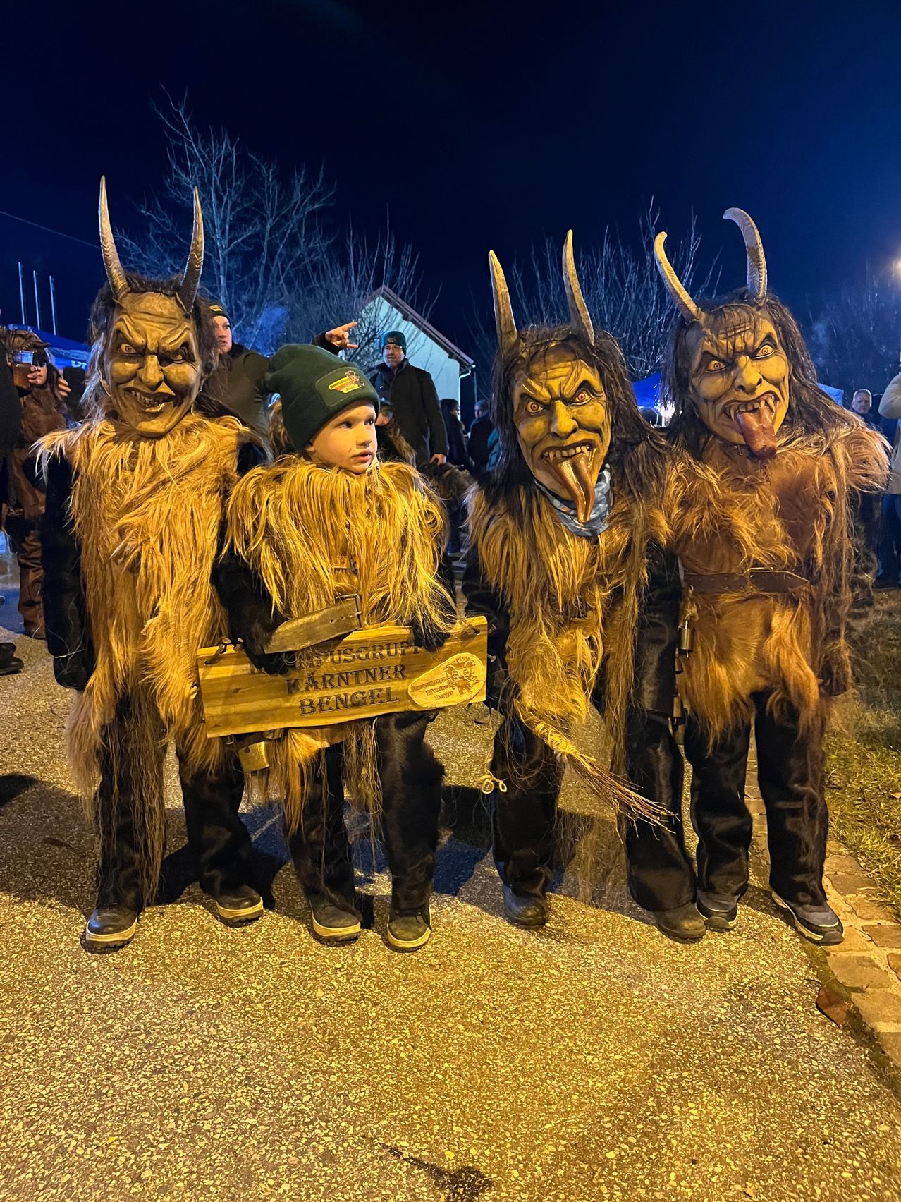 Vier Personen in gehörnten Kostümen stehen bei Nacht zusammen, ein Kind hält ein Schild mit der Aufschrift 'Kärntner Bengel'. Die Atmosphäre ist festlich mit einem Hintergrund aus Bäumen und Menschen.