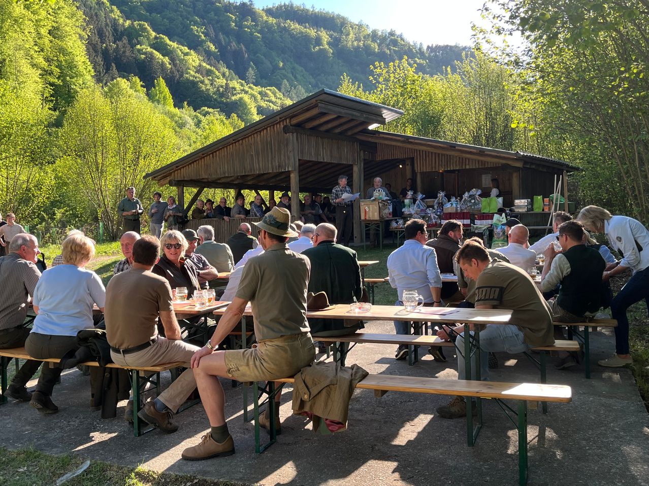 In einer Freiluftumgebung sitzen Menschen an Tischen, einige tragen Hüte und Stiefel. Im Hintergrund befindet sich ein hölzernes Schutzdach, um das herum Menschen stehen. In der Ferne sind Berge zu sehen.