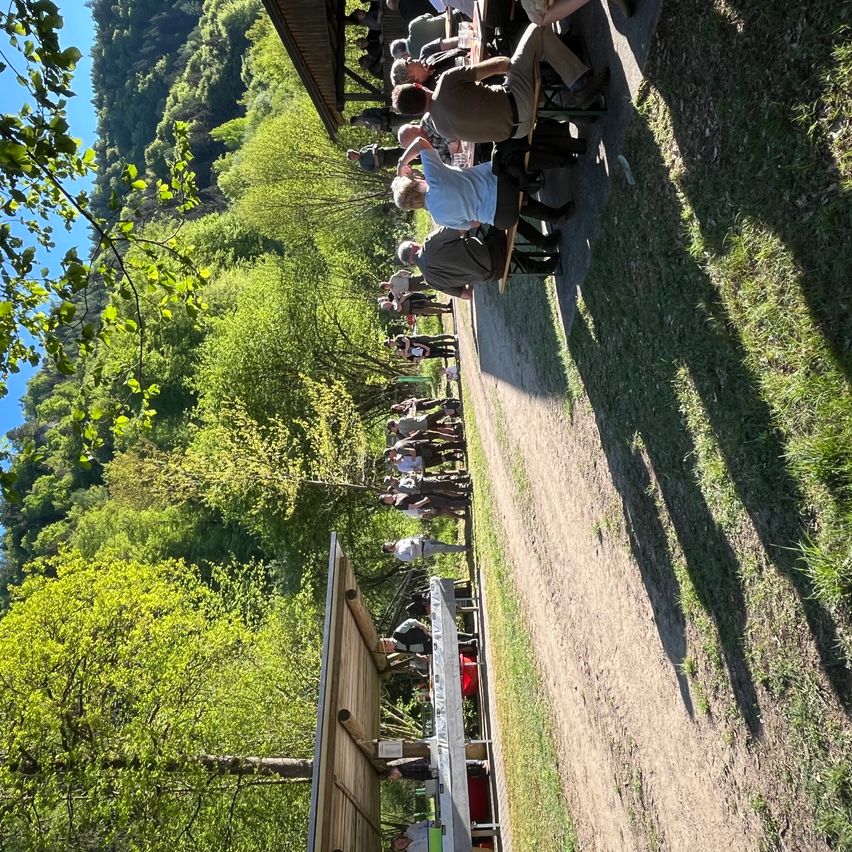 Eine Gruppe von Menschen sitzt an einem langen Picknicktisch an einem sonnigen Tag, umgeben von Bäumen.