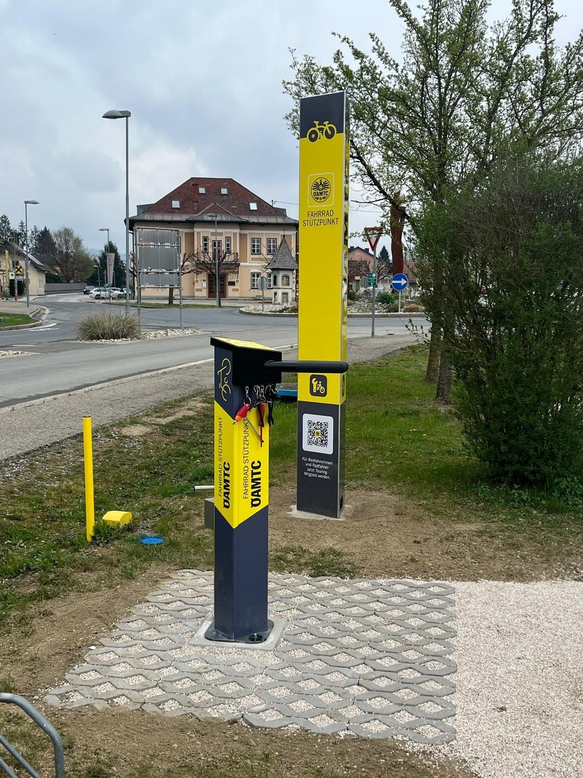 Ein gelber und schwarzer Fahrradständer mit QR-Code steht auf einem gepflasterten Bereich mit einem Gebäude im Hintergrund.