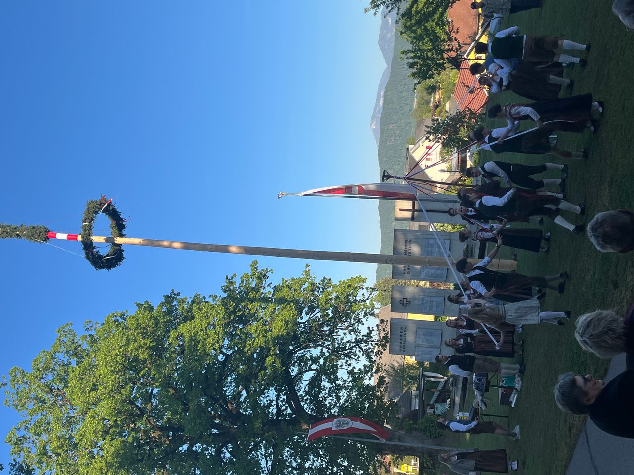 Eine Gruppe von Menschen in traditionellen Kostümen führt an einem sonnigen Tag ein Ritual mit Flaggen und einem mit einem Kranz geschmückten Pfahl durch.