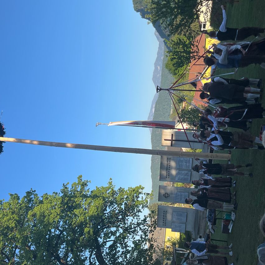 Eine Gruppe von Menschen in traditioneller Kleidung versammelt sich in einem Park und steht um einen hohen Fahnenmast. Sie scheinen ein traditionelles Fest zu feiern.