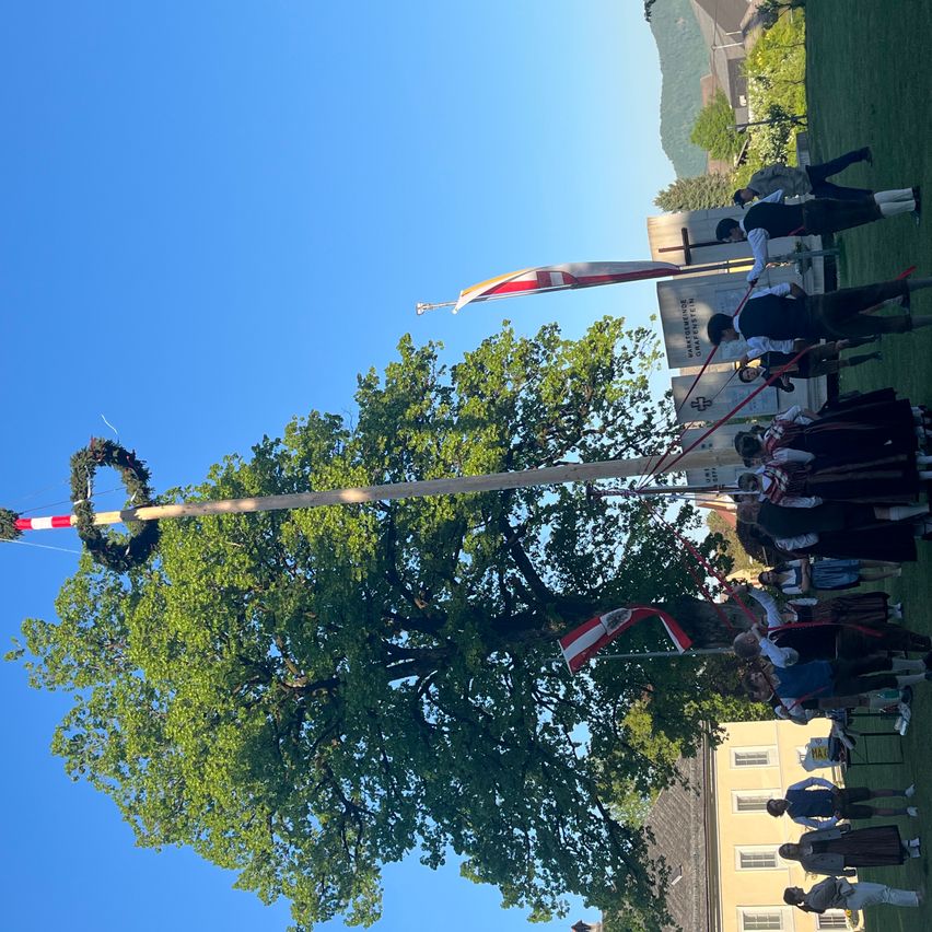 Ein großer Baum ist mit einem Kranz und einer Flagge geschmückt. Leute stehen im Kreis darum. Einige tragen traditionelle Kleidung. Hinter dem Baum befinden sich einige Grabsteine und ein Gebäude.