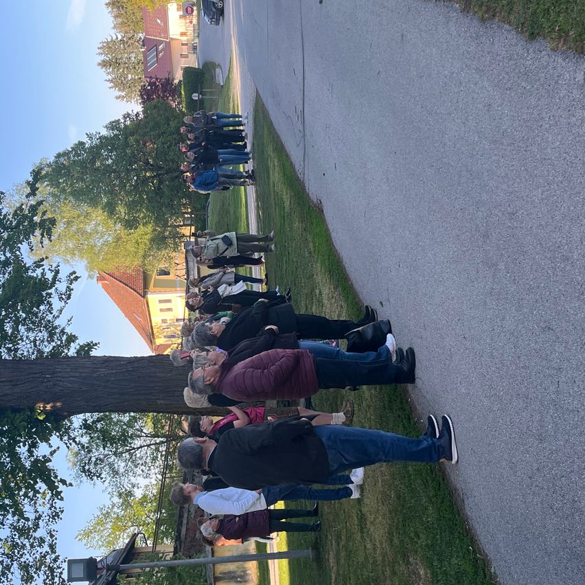 Eine Gruppe von Menschen steht in einer Reihe auf einem Bürgersteig, einige tragen Turnschuhe und andere Stiefel, mit einem großen Baum und einem gelben Gebäude im Hintergrund.