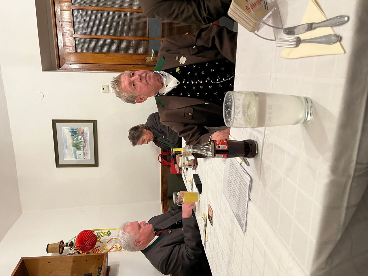 Zwei Männer sitzen an einem Tisch, einer mit einem Glas Bier, der andere mit einem Glas Wasser. Sie befinden sich in einem Raum mit einer Holztür, einem Bilderrahmen und einer gefliesten Wand.