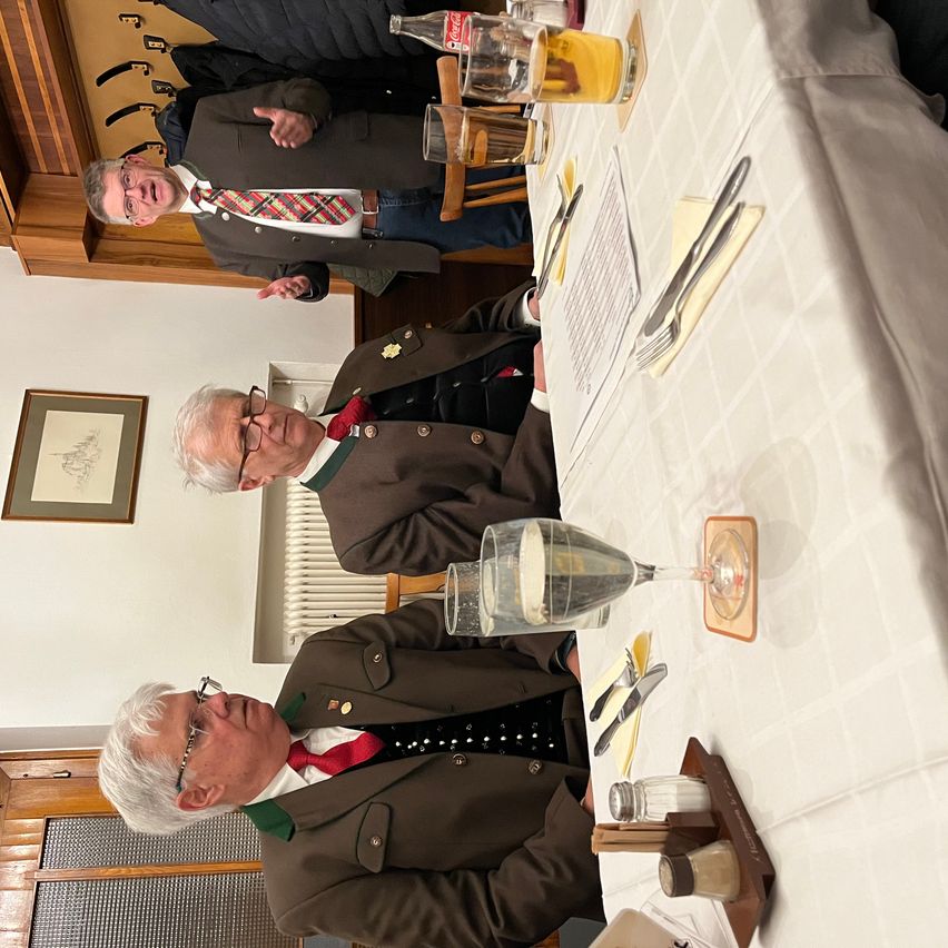 Drei Männer in formeller Kleidung, zwei sitzen an einem Tisch mit Essen und Getränken, einer steht hinter ihnen in einem Raum.