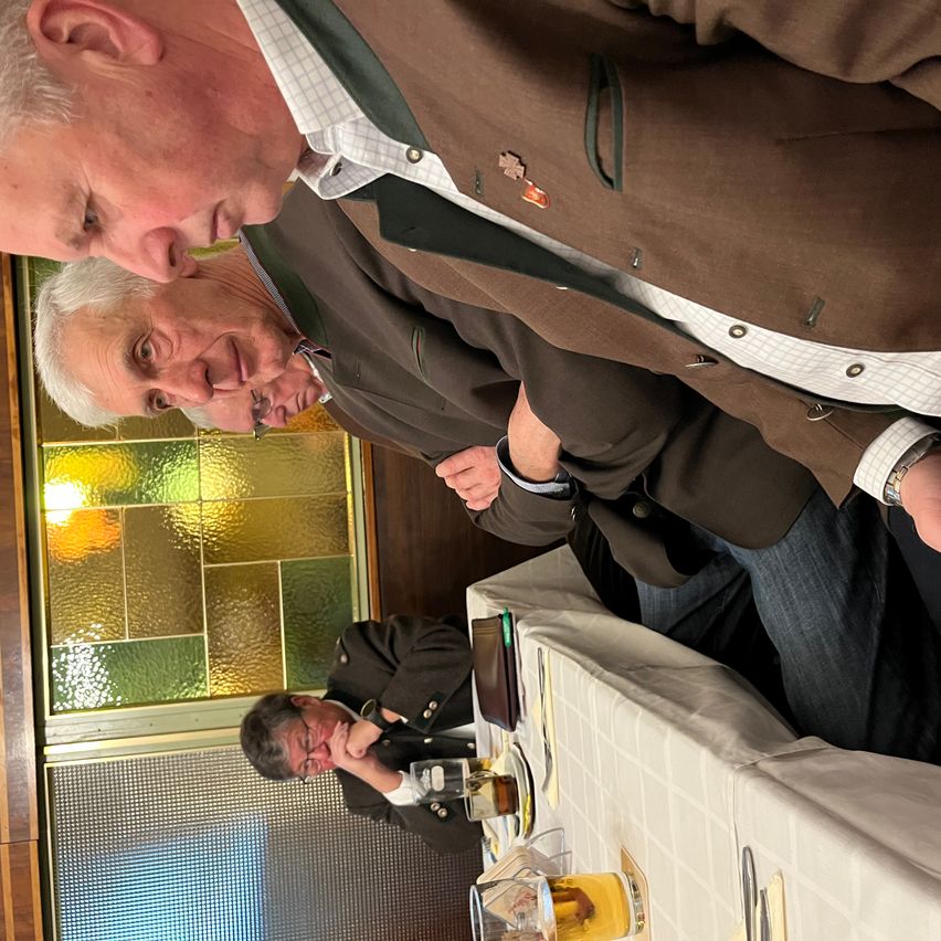 Drei Männer in Anzügen sitzen um einen Tisch mit Getränken, einer lehnt sich vor, um mit einem anderen zu sprechen. Hinter ihnen erhellt ein farbenfrohes Buntglasfenster die Szene.