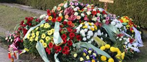 Eine große Ansammlung von Blumen, darunter Rosen, Gänseblümchen und Lilien, ist in einem Halbkreis um ein kleines Holzkreuz angeordnet.