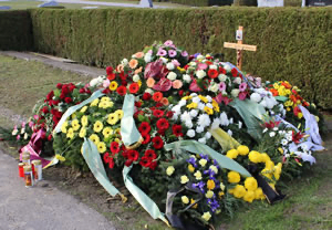 Eine große Ansammlung von Blumen, darunter Rosen, Gänseblümchen und Lilien, ist in einem Halbkreis um ein kleines Holzkreuz angeordnet.