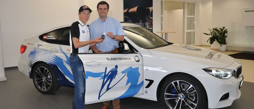 Zwei Männer stehen neben einem BMW M4 Coupé. Einer hält ein kleines Objekt, während der andere eine Kappe trägt. Das Auto hat ein blau-weißes Design.