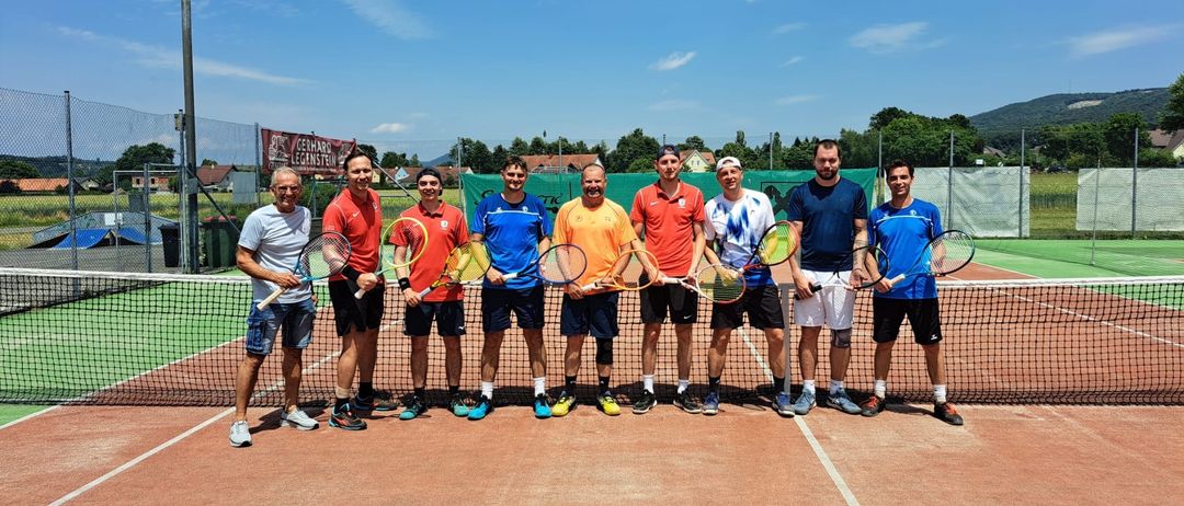 Eine Gruppe von neun Männern, alle in Tenniskleidung gekleidet, steht auf einem Tennisplatz. Sie halten alle Tennisschläger. Hinter ihnen befindet sich ein Netz und ein Zaun.