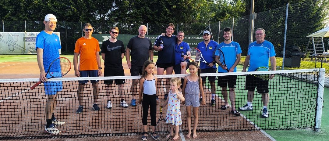 Eine Gruppe von Männern und Kindern steht auf einem Tennisplatz, hält Schläger und zwei Mädchen in der Mitte lächeln und posieren für ein Foto.