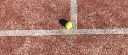 Ein Tennisball liegt auf einem Tennisplatz mit braunen und weißen Markierungen. Der Platz ist leer.