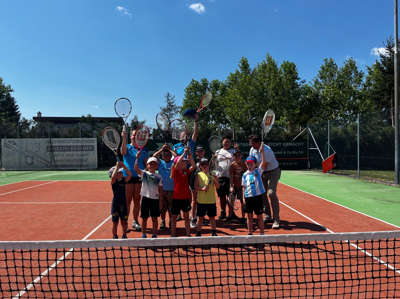 Eine Gruppe von Kindern und Erwachsenen steht auf einem Tennisplatz, hält Tennisschläger und posiert für ein Foto. Der Platz ist von einem Zaun mit einem Banner umgeben.