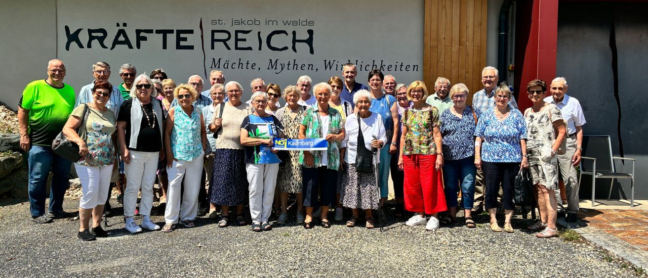 Eine Gruppe von Menschen steht vor einem Gebäude mit 'St. Jakob im Walde' und 'Fete Reich' darauf geschrieben. Sie tragen Freizeitkleidung und einige halten ein Schild mit 'NEIN' und 'Kaumberg.'