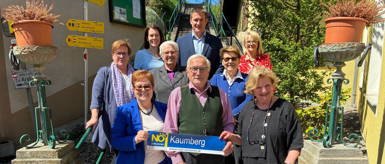 Eine Gruppe von Menschen steht auf einer Treppe mit einem blauen Banner, auf dem 'NO Kaumberg' steht. Sie tragen Freizeitkleidung und Brillen. Eine Person hält ein blaues Banner mit weißem Text und einem Logo. Hinter ihnen stehen Pflanzen und Schilder an einem Pfosten.