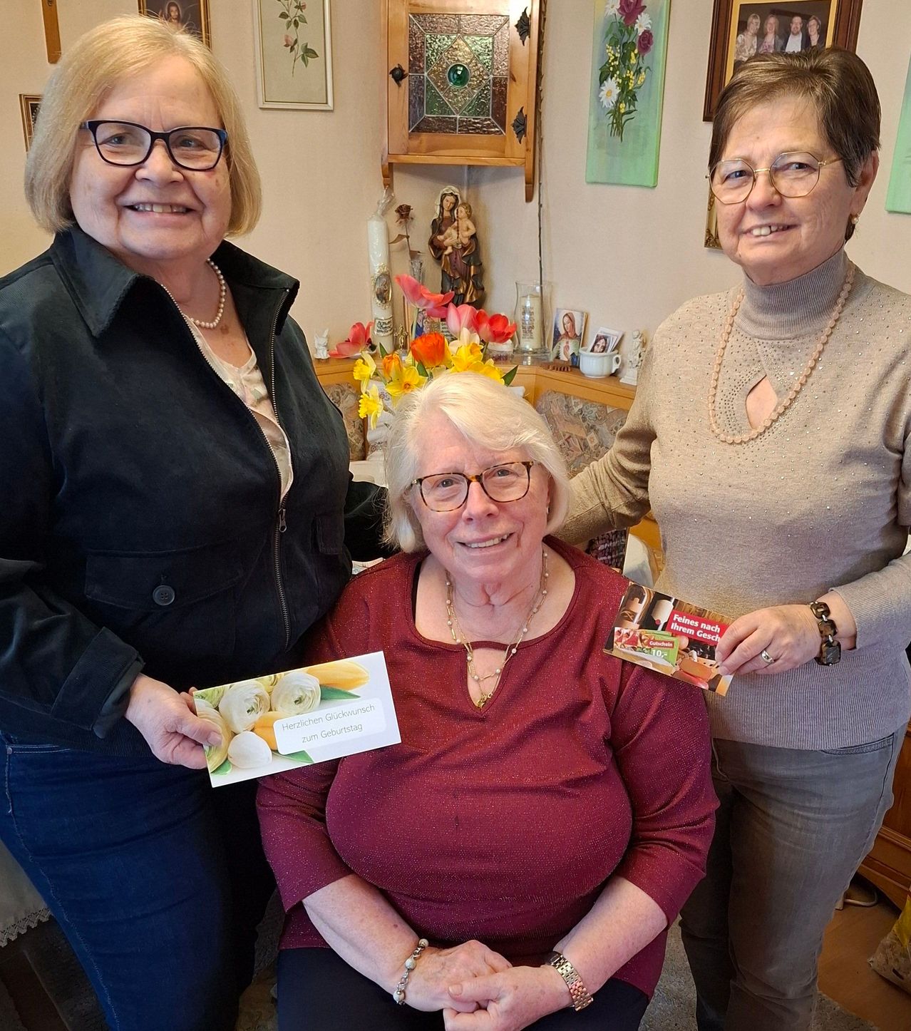Three women in a living room, one holding a birthday card for another woman, all smiling and posing for a photo.