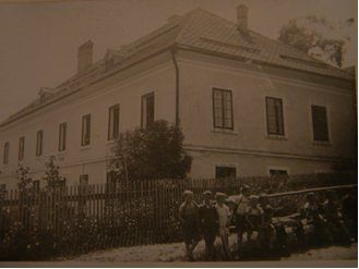 Ein altes Foto zeigt eine Gruppe von Menschen vor einem großen, weißen Gebäude mit mehreren Fenstern und einem Ziegeldach. Sie stehen vor einem Holzzaun.