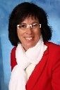 Eine Frau mit Brille und rotem Blazer lächelt für ein Foto. Sie hat kurzes schwarzes Haar und trägt einen weißen Schal. Der Hintergrund ist blau.