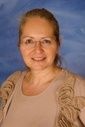 Eine lächelnde Frau mit kurzen Haaren und Brille steht vor einem blauen Hintergrund. Sie trägt ein beiges Oberteil und hat einen warmen Ausdruck im Gesicht.