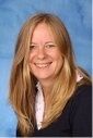 Eine Frau mit langen blonden Haaren lächelt und posiert für ein Foto. Sie trägt ein schwarzes Oberteil und ein weißes Hemd mit Kragen. Der Hintergrund ist eine einfarbige blaue Farbe.