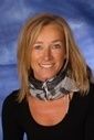 Eine Frau mit blondem Haar lächelt und trägt ein schwarzes Oberteil und einen Schal. Sie steht vor einem blauen Hintergrund.