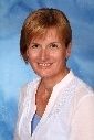 Eine lächelnde Frau mit kurzen blonden Haaren, die ein weißes Oberteil trägt, steht vor einem blauen Hintergrund.