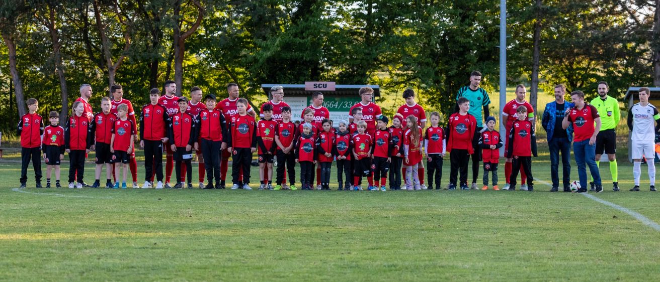 Eine Gruppe junger Fußballspieler, zusammen mit Trainern, steht auf einem Feld und posiert wahrscheinlich für ein Teamfoto. Sie tragen rote Uniformen mit Nummern, und der Hintergrund zeigt Bäume und ein Gebäude.