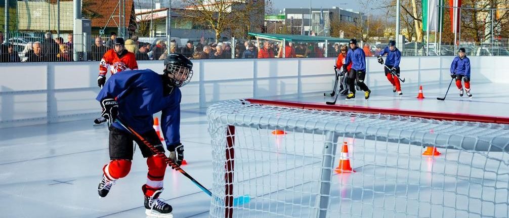 Eine Gruppe von Eishockeyspielern auf einer Eisbahn, die Helme, Handschuhe und Schlittschuhe tragen, skatet in verschiedene Richtungen. Es gibt orangefarbene Kegel und Straßenlaternen auf der Eisbahn.