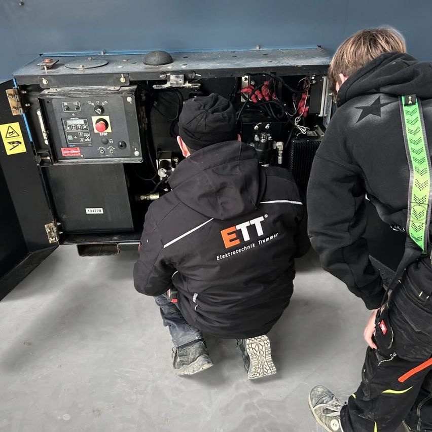 Two people in black jackets with ETT logo are working on a machine with wires and a red button in a room.