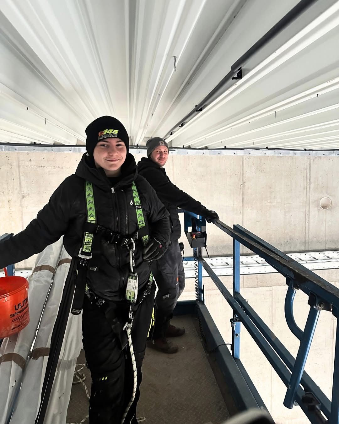 Two workers in winter gear stand on a metal scaffolding. One holds a bucket, both wear safety harnesses. They are under a concrete ceiling with metal beams.