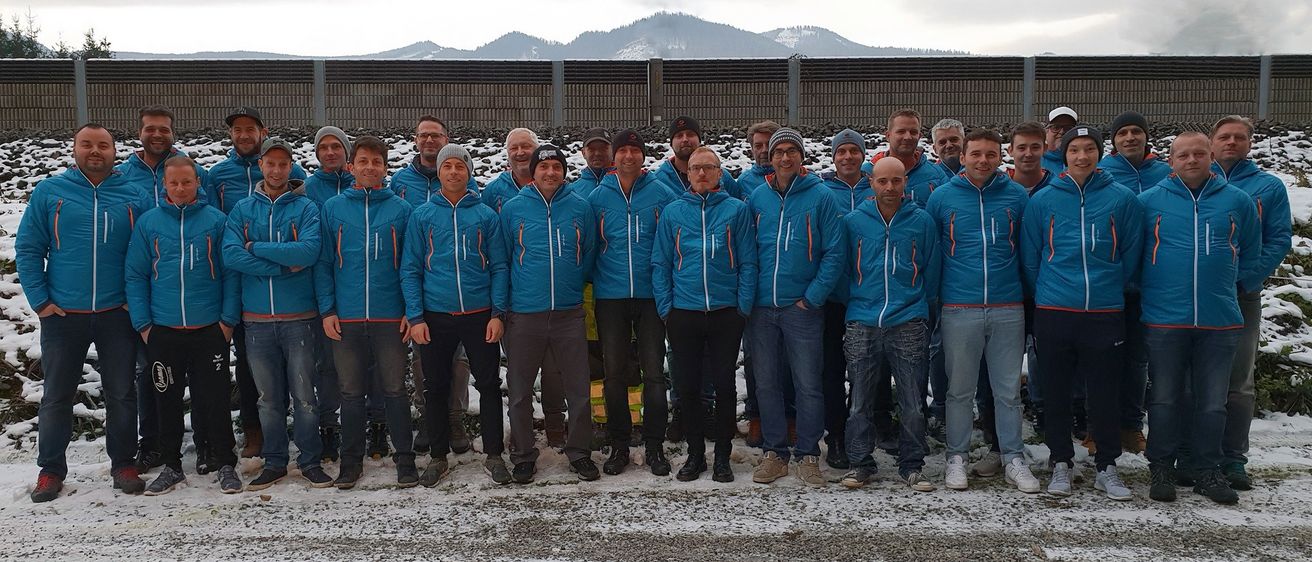 Eine Gruppe von Männern in blauen Jacken steht auf einem verschneiten Boden mit Bergen im Hintergrund. Einige tragen Brillen und Mützen. Sie scheinen für ein Foto zu posieren.
