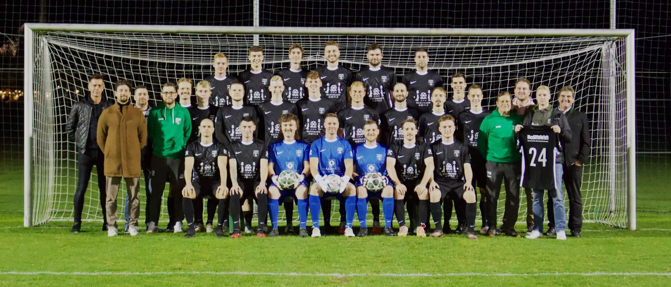 Eine Fußballmannschaft posiert für ein Foto auf einem Feld, Spieler in schwarzen und blauen Trikots, einige halten Fußballbälle. Ein Tornetz ist hinter ihnen.