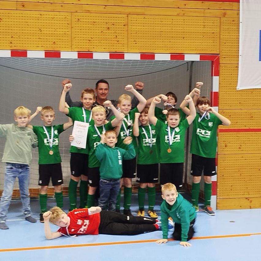 Eine Gruppe junger Jungen in grünen Trikots und Medaillen feiert einen Sieg in einer Sporthalle, posiert mit erhobenen Fäusten und lächelt. Ein Mann steht hinter ihnen.