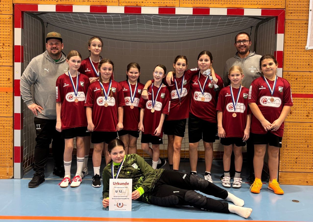 Eine Gruppe junger Mädchen in roten Trikots und Medaillen posiert vor einem Tor. Ein Mädchen liegt auf dem Boden und hält ein Zertifikat mit dem Text 'U12'. Zwei Männer stehen hinter ihnen.