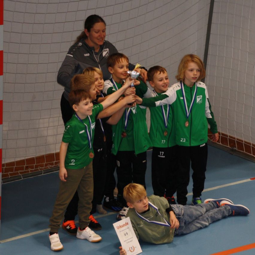Eine Gruppe junger Athleten in grünen Trikots feiert ihren Sieg mit einem Pokal, Medaillen und einem Zertifikat in einer Sporthalle. Eine Frau steht lächelnd hinter ihnen.