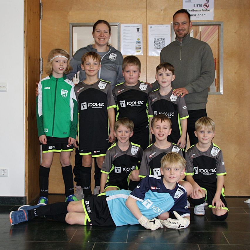 Eine Gruppe junger Fußballer, die schwarze, graue und grüne Trikots tragen, posiert für ein Foto. Hinter den Kindern stehen zwei Erwachsene, ein Mann und eine Frau. Die Kinder tragen Handschuhe, und einige von ihnen tragen Stirnbänder. Das Bild hat einen Wasserzeichen in der oberen rechten Ecke.