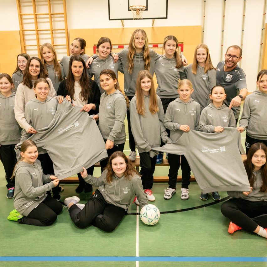 Eine Gruppe junger Mädchen in grauen Uniformen posiert für ein Foto in einer Turnhalle mit einem Fußball auf dem Boden.
