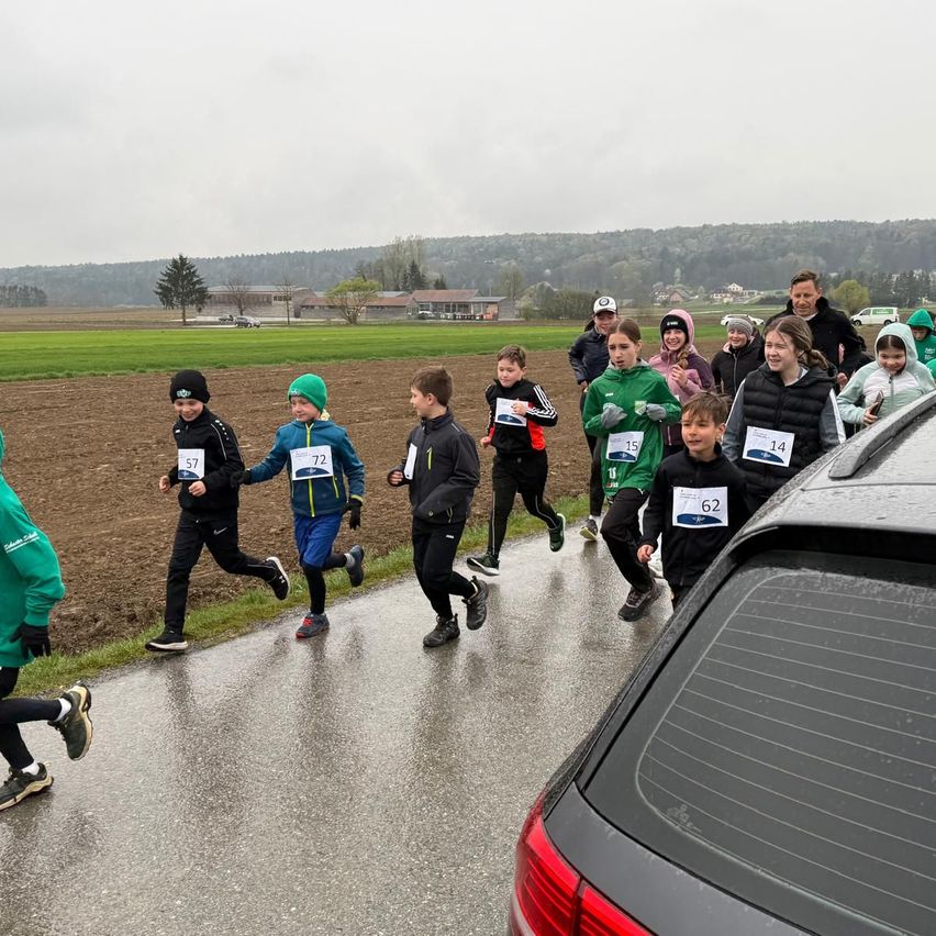 A group of young runners participate in a race on a wet road with a car parked nearby. Behind them, adults and more children follow. The backdrop includes a field and distant hills.