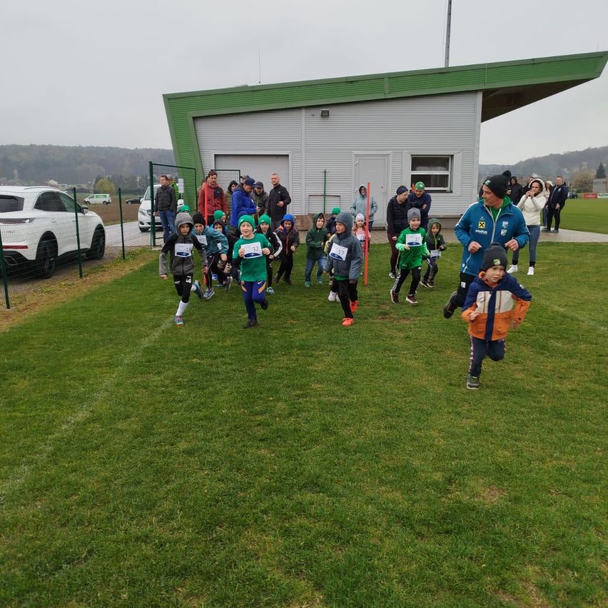 Children participate in a race on a grassy field with a building in the background. Spectators watch from behind a fence.