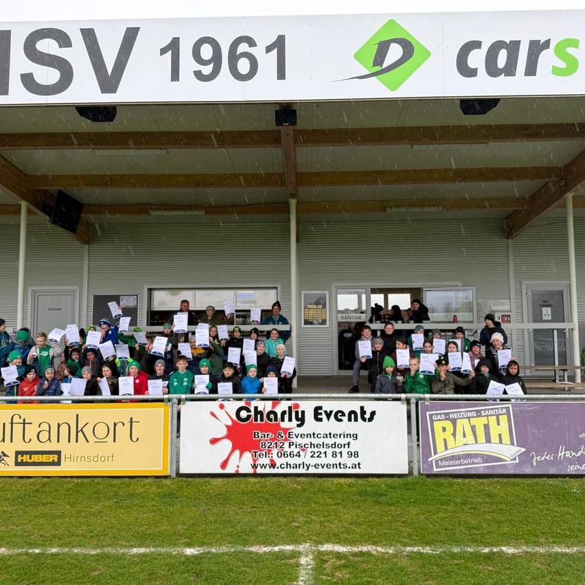 A group of children in green shirts and hats pose for a photo under a sign that reads D cars, 1961. They are on a field with a fence, banners, and a canopy.