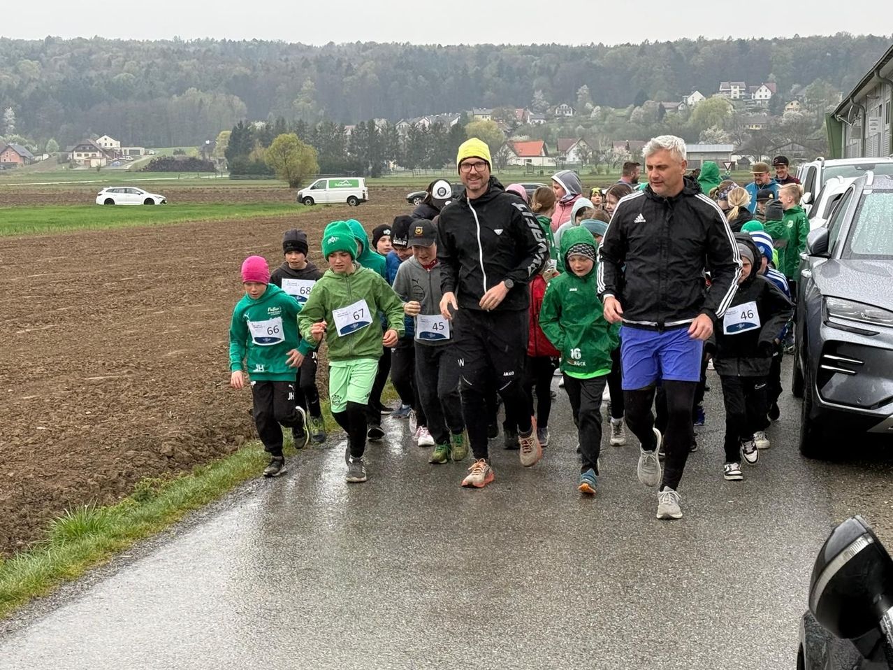 A group of children and adults are running in a race. They wear green shirts with numbers. Two men are walking beside them. Behind them is a field with houses and trees.