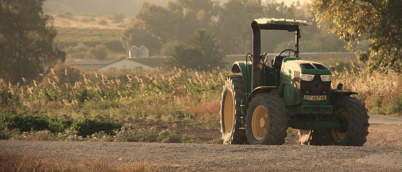 Ein grüner Traktor mit gelben Felgen steht auf einem Schotterweg in einem ländlichen Gebiet mit einem Berg und Bäumen im Hintergrund.