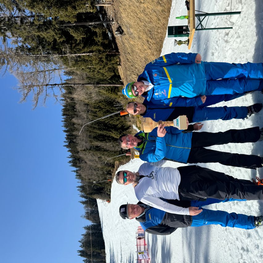Vier Männer in Skiausrüstung posieren auf einem verschneiten Hügel mit Kiefernbäumen und einer hölzernen Struktur im Hintergrund. Sie halten Pokale und scheinen zu feiern.